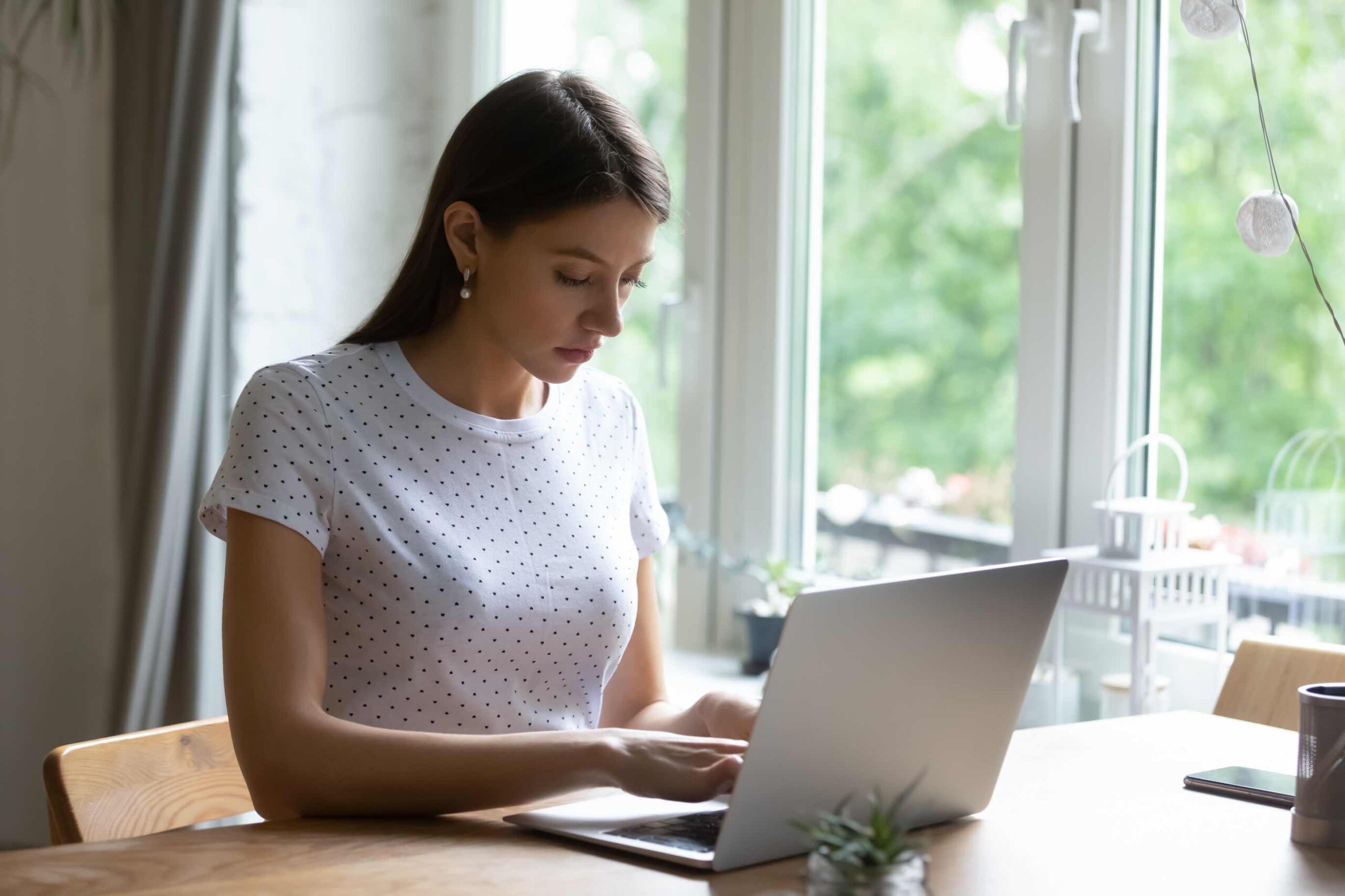 A woman typing on a laptop inside by a window.
